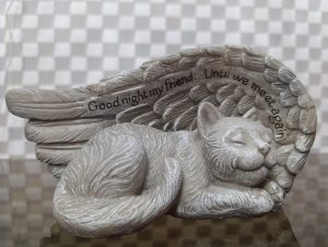 A decorative figurine of a sleeping cat, crafted from a light-colored material. The cat is nestled against a set of angelic wings that envelop it protectively.
