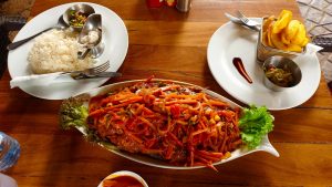 A wooden table featuring a large, oval platter of whole fried fish topped with a thick vegetable sauce of sliced carrots and onions. Accompanying plates hold a portion of white rice with small sauce boats and a wire basket of golden potato chips. A water bottle and small dipping sauce bowl are also visible on the table surface.