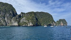A wide view of massive, lush green limestone islands in the ocean, surrounded by several tourist boats and speedboats in the clear blue water.