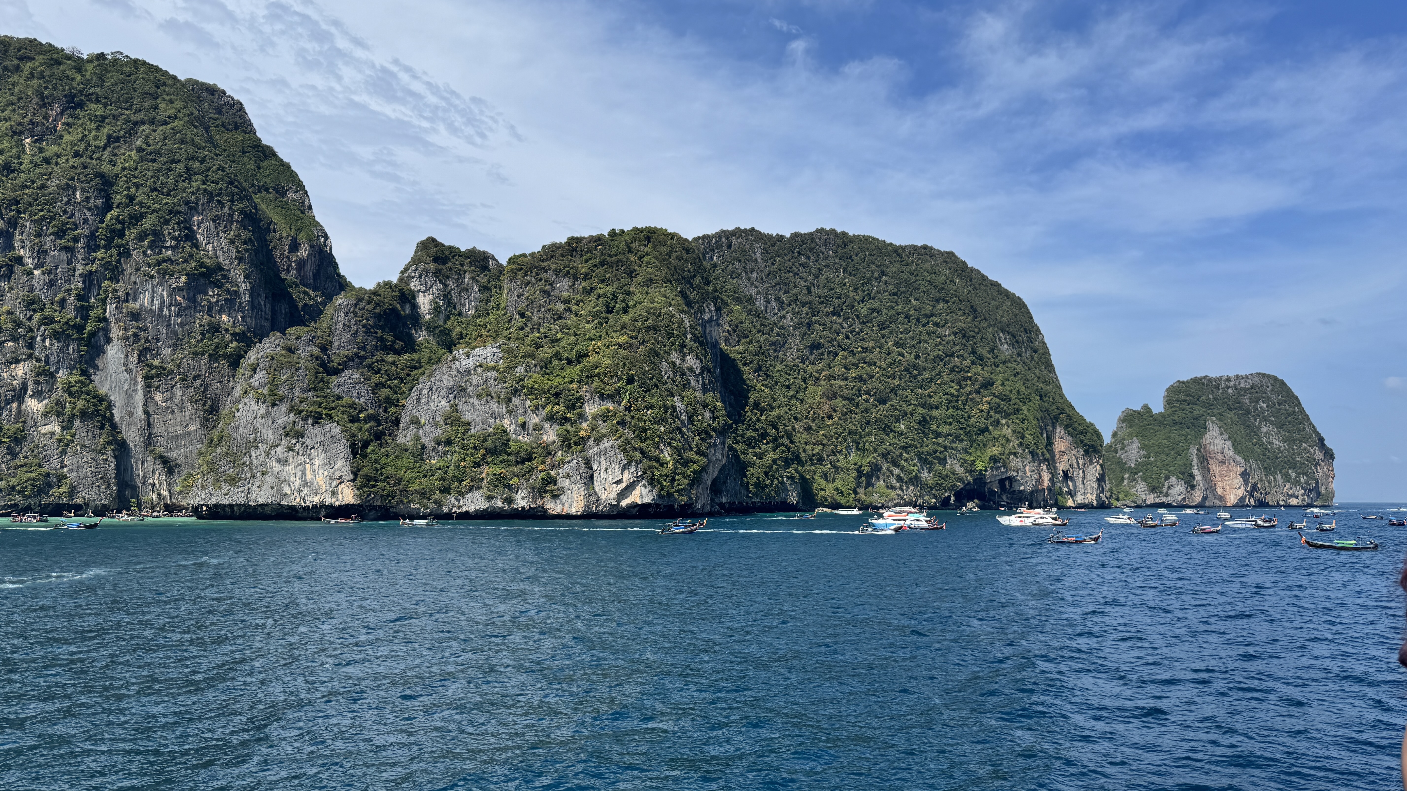 A wide view of massive, lush green limestone islands in the ocean, surrounded by several tourist boats and speedboats in the clear blue water.