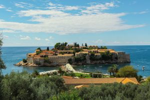 A picturesque islet packed with charming stone buildings and red-tiled roofs is connected to the mainland by a narrow stone walkway. Surrounded by the deep blue waters of the Adriatic Sea, the scene is framed by lush green trees and a soft, cloudy sky.

