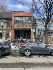 An old brick two-storey shop in Toronto’s Kensington Market neighbourhood fronts a wide sign with block lettering that reads “HARDBOILED,” while a smaller, square  sign in front of it affixed to a utility pole reads “A LITTLE DARKNESS IS OKAY” in a stylized script font. Blue and gray cars are parked at the curb in front of the building.
