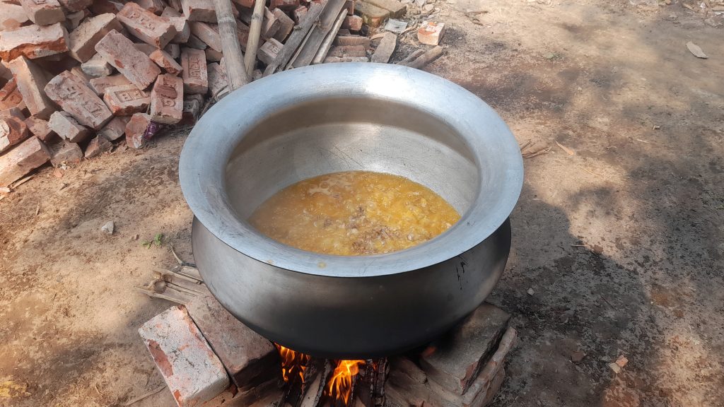 A large metal pot with bubbling stew sits on an outdoor brick fireplace, with bricks and logs nearby.