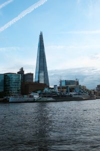 A vertical photo of the London skyline across the river, featuring a tall glass skyscraper and a historic ship in the foreground.