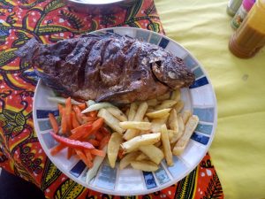 A top-down view of a whole fried tilapia with chips and a fresh vegetable salad on a patterned plate, set on a bright orange-yellow tablecloth with a napkin and condiments nearby.
