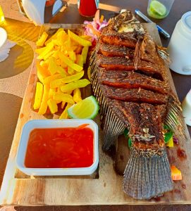 Whole deep-fried tilapia with fries, chili sauce, lime, and kachumbari served on a wooden board.