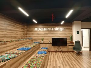 An interior shot of a lounge area featuring wooden-paneled walls and flooring. A tiered wooden seating structure is lined with colorful patterned cushion with African prints. A television on a tripod stand, a green armchair, and several potted plants complete the space.