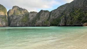 A serene tropical bay with turquoise waters, white sandy beach, and limestone cliffs lush with green trees under a cloudy sky.
