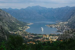A large cruise ship glides through a narrow, winding bay surrounded by charming coastal towns with red-roofed buildings. Towering, rocky mountains rise steeply from the water’s edge, creating a breathtaking fjord-like landscape under a soft sky.
