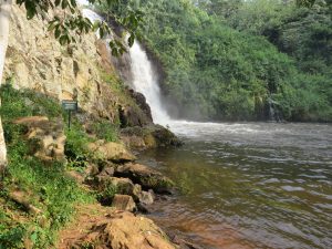Small waterfalls in Uganda flow down a rocky cliff into a pool, surrounded by dense tropical greenery, with a warning sign and rippling water in the foreground.

