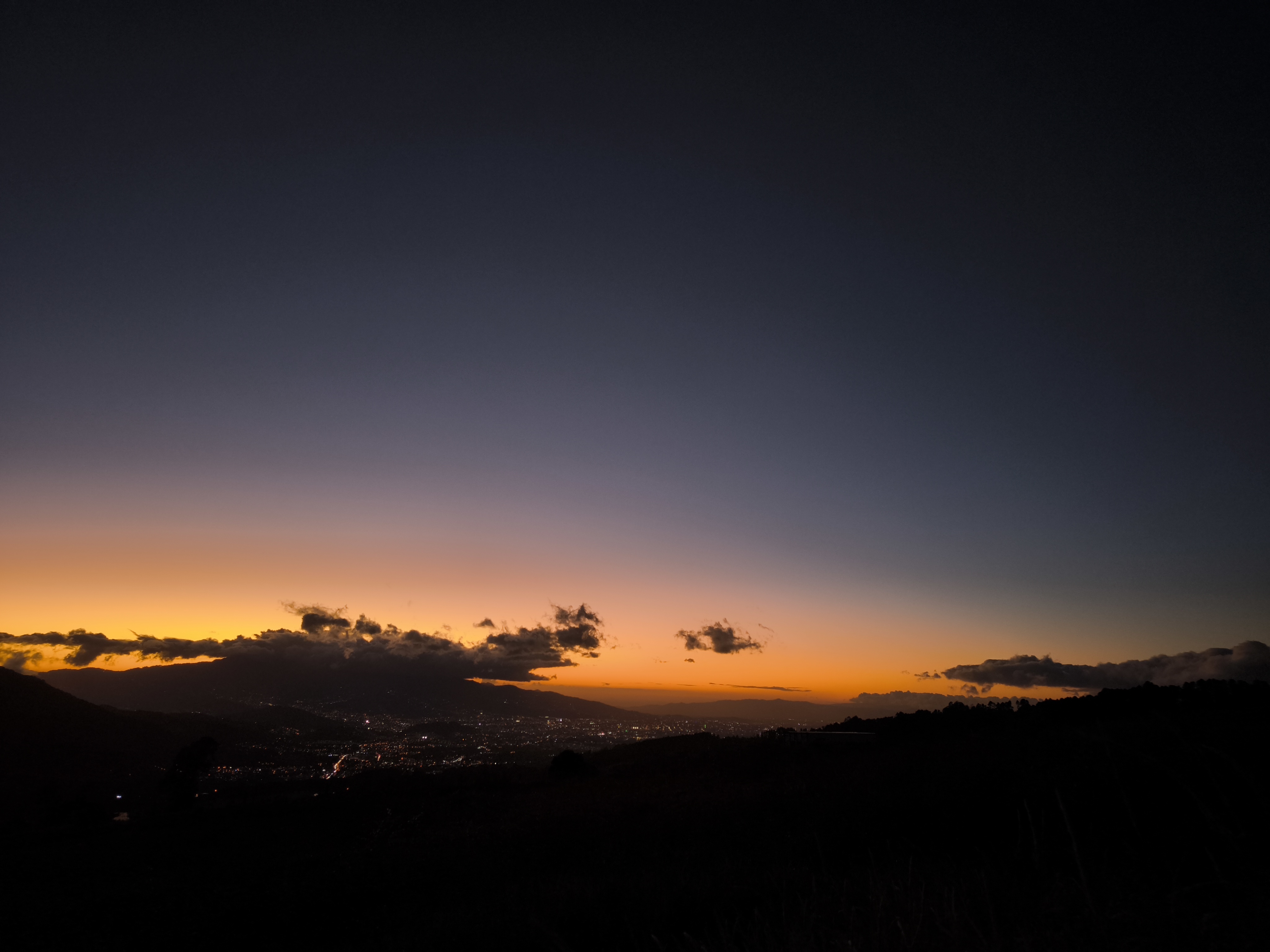 A twilight view over a city valley with an orange horizon fading into deep blue, clouds above mountains, and city lights below.