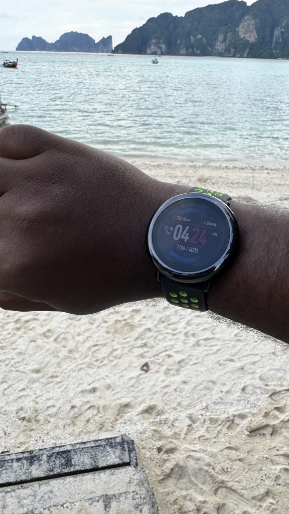 A close-up of a wrist wearing a black smartwatch with a green and black strap. The background shows a sandy beach and the ocean.