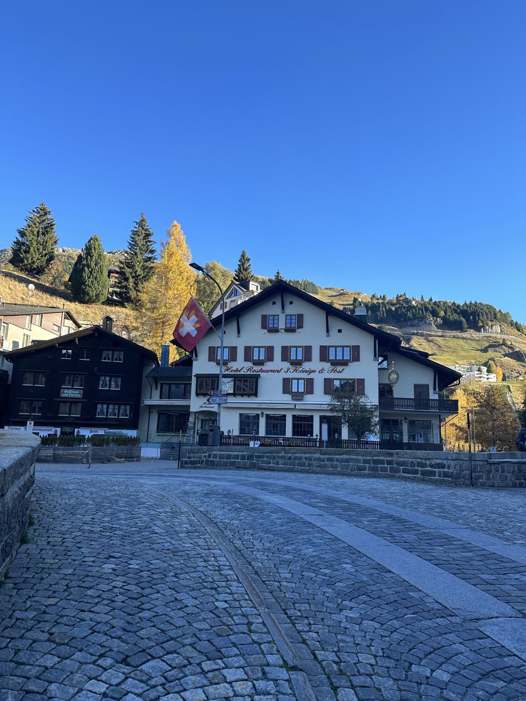 A picturesque scene featuring a cobblestone street lined with traditional Swiss buildings. In the foreground, a curved stone pathway leads towards a white building with dark wooden accents and large windows, displaying a sign for a restaurant. The building features a Swiss flag prominently waving in front. Surrounding trees, some in autumn gold, add a touch of nature, while a clear blue sky enhances the serene atmosphere of the alpine village. The distant hills are visible in the background, completing the scenic view.