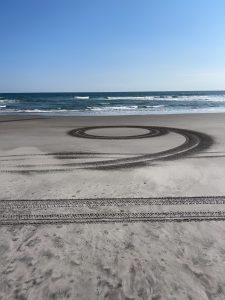 
A sandy beach with gentle waves rolling in from the ocean. In the foreground, there are circular tire tracks imprinted in the sand, creating a spiral pattern
