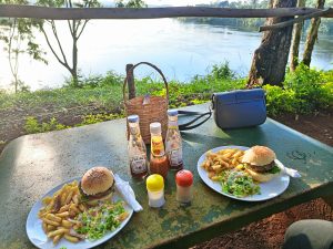Two plates of burgers with fries and side salads sit on a rustic table by a calm river at sunset, with condiments and cutlery nearby.