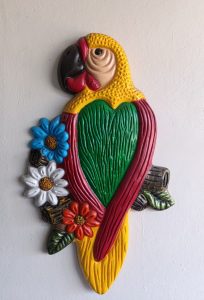 A colorful decorative wall hanging shaped like a parrot, featuring vibrant colors including yellow, green, red, and black. The parrot is perched among stylized flowers in blue, white, and orange, with green leaves and wooden accents, all set against a light-colored wall.