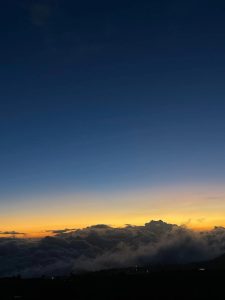 A serene sunset over a sea of clouds, gradually transitioning from deep blue at the top to warm oranges and yellows near the horizon.
