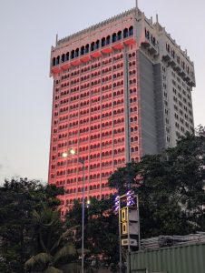 A tall, grand hotel tower featuring arched windows illuminated with a vibrant red glow, set behind lush green trees under a pale evening sky.
