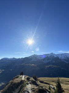 A panoramic view of a mountainous landscape under a clear blue sky. The sun is shining brightly in the upper center of the image, casting rays that illuminate the scene. Snow-capped peaks are visible in the distance, surrounded by rugged hills and green valleys. In the foreground, there is a grassy area with a rocky path leading towards a small group of trees and remnants of a structure. The overall atmosphere conveys a sense of tranquility and natural beauty.