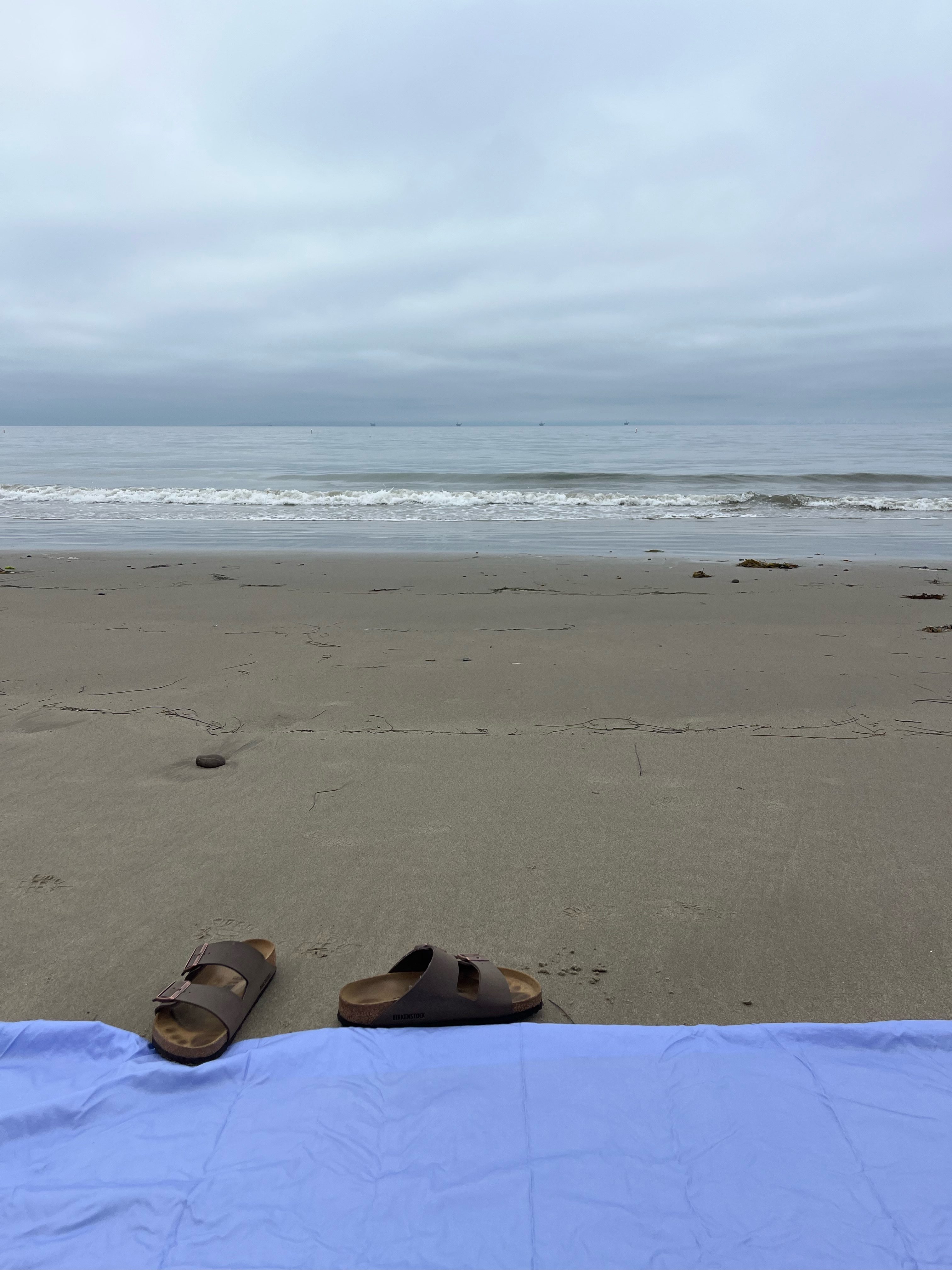 A view of a sandy beach with a calm ocean extending to the horizon under a cloudy sky. In the foreground, a light purple beach towel is laid out on the sand, next to a pair of brown sandals