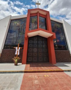 The image shows the exterior of a modern church with a prominent entrance featuring large, black doors. Above the entrance, there is a red architectural element with a cross at the top. The facade includes colorful stained glass windows depicting religious scenes. In front of the church, there is a wooden cross draped with a white cloth, accompanied by a small planter with plants. The sky is partly cloudy, adding to the ambiance of the scene.