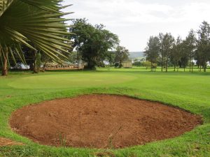 Circular sand bunker on a golf course with a green, trees, a building, and a distant lake under an overcast sky.