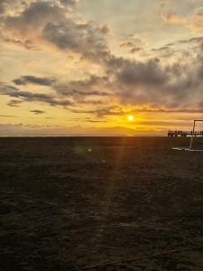 Sunset over the beach with warm golden light and a pier in the distance, creating a peaceful coastal scene.