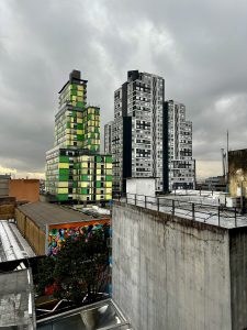 A rooftop view over central Bogotá, Colombia, under a heavy overcast sky. A striking residential tower with green and yellow mosaic panels dominates the left, flanked by grey concrete high-rises. A vibrant tropical street mural with birds and geometric shapes brightens the lower foreground.