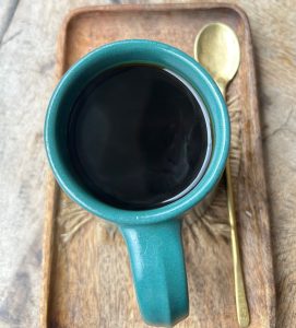 A turquoise coffee cup rests atop a wooden tray, containing dark black coffee that reflects light at its surface.
