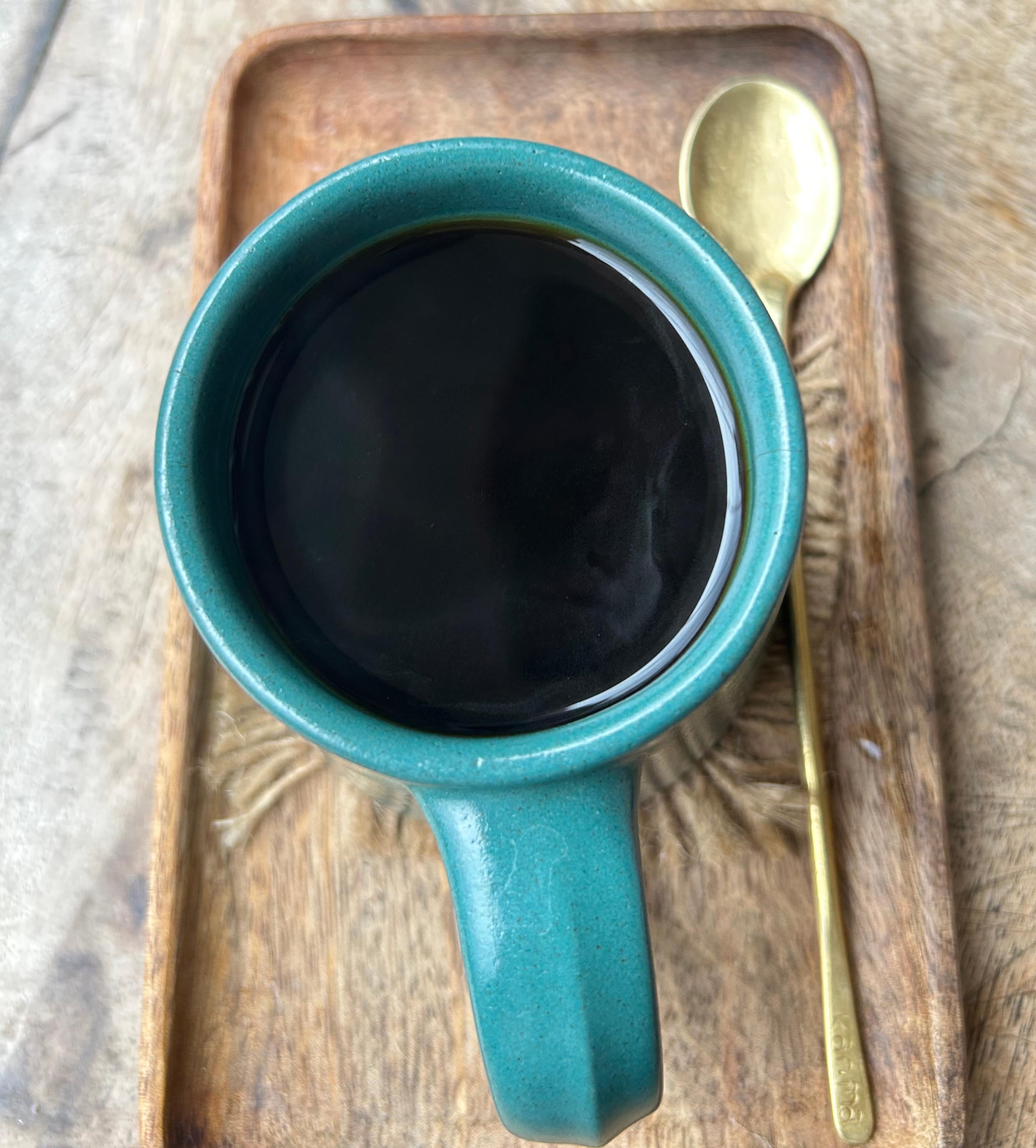 A turquoise coffee cup rests atop a wooden tray, containing dark black coffee that reflects light at its surface.