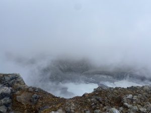 
A rocky landscape shrouded in thick fog, with a partially obscured view of a volcanic crater below.