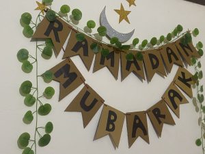 Decorative "Ramadan Mubarak" banner with green leaf garland, golden stars, and a silver crescent moon on a white wall, conveying festive cheer.