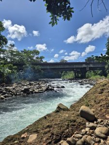 A flowing river beneath a bridge under a clear blue sky with scattered clouds, surrounded by rocks and lush greenery along the banks
