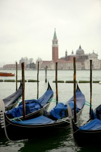 Gondolas tied to wooden poles on calm water, with a historic building and bell tower under an overcast sky.