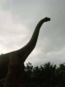 A silhouette of a long-necked dinosaur sculpture against a cloudy sky, surrounded by lush greenery. The dinosaur's neck stretches upward, emphasizing its height and large stature. 