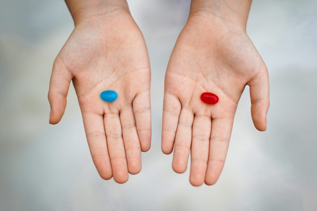 Two open hands are shown, each holding a pill. The left hand holds a blue capsule, while the right hand holds a red capsule. The background is softly blurred, emphasizing the contrasting colors of the pills against the skin of the hands.