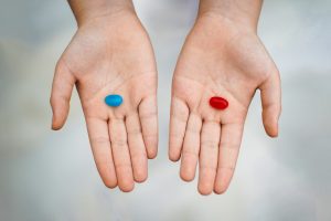 Two open hands are shown, each holding a pill. The left hand holds a blue capsule, while the right hand holds a red capsule. The background is softly blurred, emphasizing the contrasting colors of the pills against the skin of the hands.

