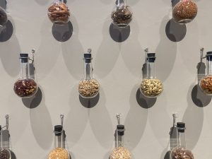 Clear glass flasks containing various spices and seeds hang on a light gray wall, casting soft shadows. The display has an organized and minimalist feel.