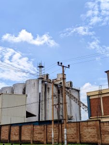 
A large industrial structure with several cylindrical silos is situated against a blue sky with scattered clouds.