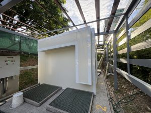 A structure with a white wall and a raised roof is seen in the image. There are two green gridded mats on a concrete platform, and next to them is a small white bucket. To the left, a control panel with buttons and lights is visible. Surrounding the structure are metal frames and a green mesh fence. In the background, greenery and a tall building are partially visible under a cloudy sky.