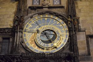 A close-up night view of the intricate Prague Astronomical Clock with golden hands and symbols on a historic stone building. Features detailed zodiac signs, figures, and Roman numerals under warm lighting.