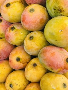 A close-up image of a pile of ripe mangoes, showcasing their vibrant yellow and pink skin. The mangoes are glossy with a fresh appearance, some displaying small dark spots and their characteristic round shape. The arrangement of the fruits creates a visually appealing and colorful display.
