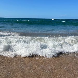 Ocean waves crash onto a sandy beach under a clear blue sky. White foam spreads across the shore as the water rolls in, creating a dynamic contrast between deep blue sea and light brown sand. The horizon stretches cleanly in the distance, evoking a calm coastal scene.