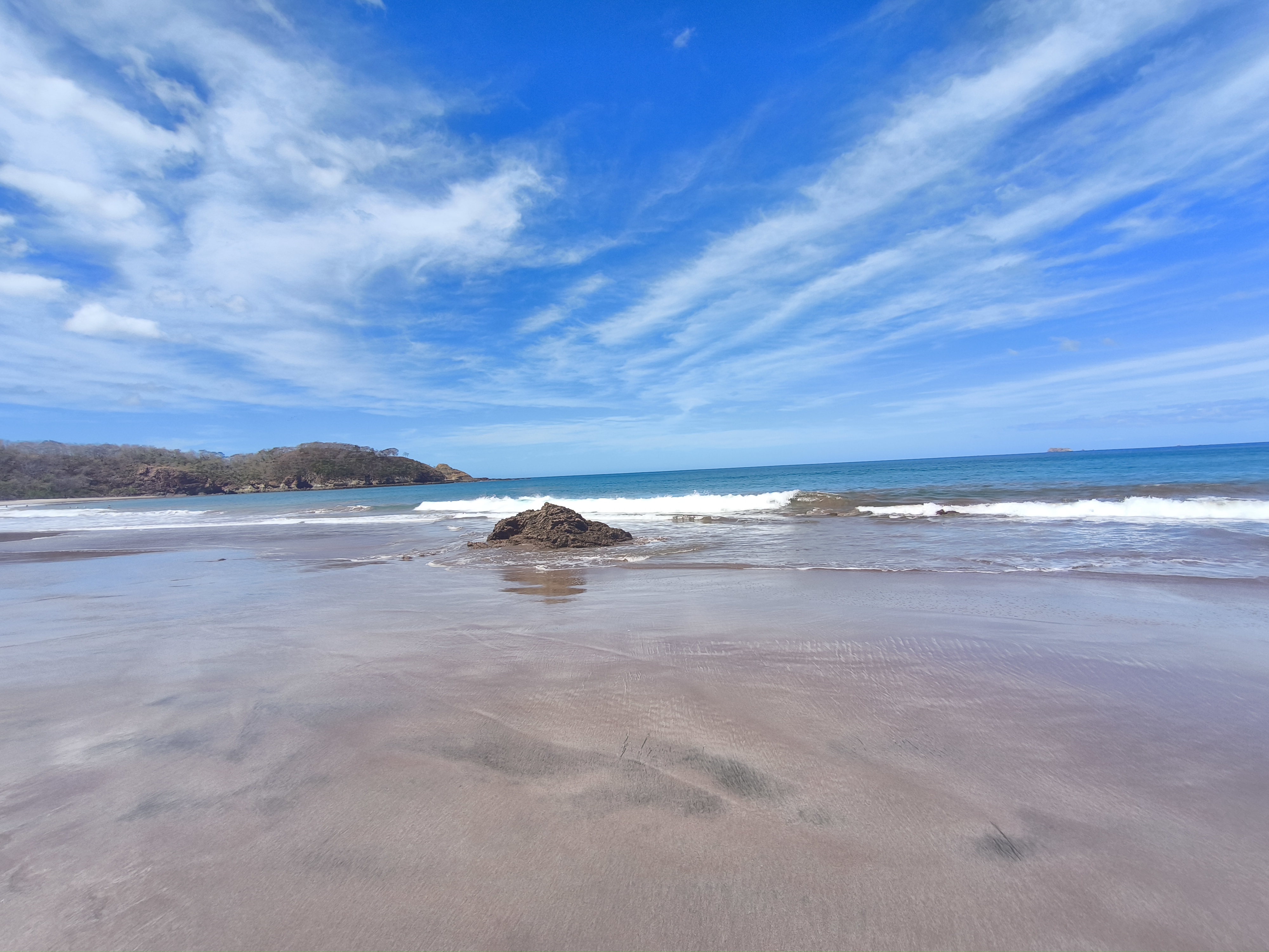 The image displays a wide, scenic coastal view of Playa El Coco in Nicaragua. Under a vibrant blue sky with wispy clouds, gentle waves roll onto the dark, shimmering volcanic sand. A small, rugged rock formation stands near the shoreline, while lush, forested hills frame the distant horizon, creating a peaceful and untouched atmosphere.