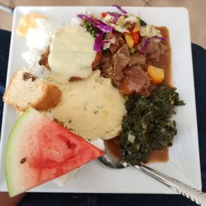 A square white plate contains Kenyan food with rice, mashed potatoes, beef stew, greens, watermelon, and a slice of cake.