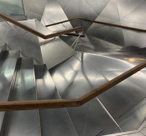 A modern staircase features sleek, metallic steps and a gracefully curved wooden handrail. Geometric wall panels surround the structure, reflecting light to create a dynamic, glowing environment that highlights the metallic surfaces.