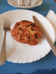 A square serving of Melanzane alla Parmigiana with layered eggplant and browned cheese on a white plate, with a fork and knife on a blue surface.