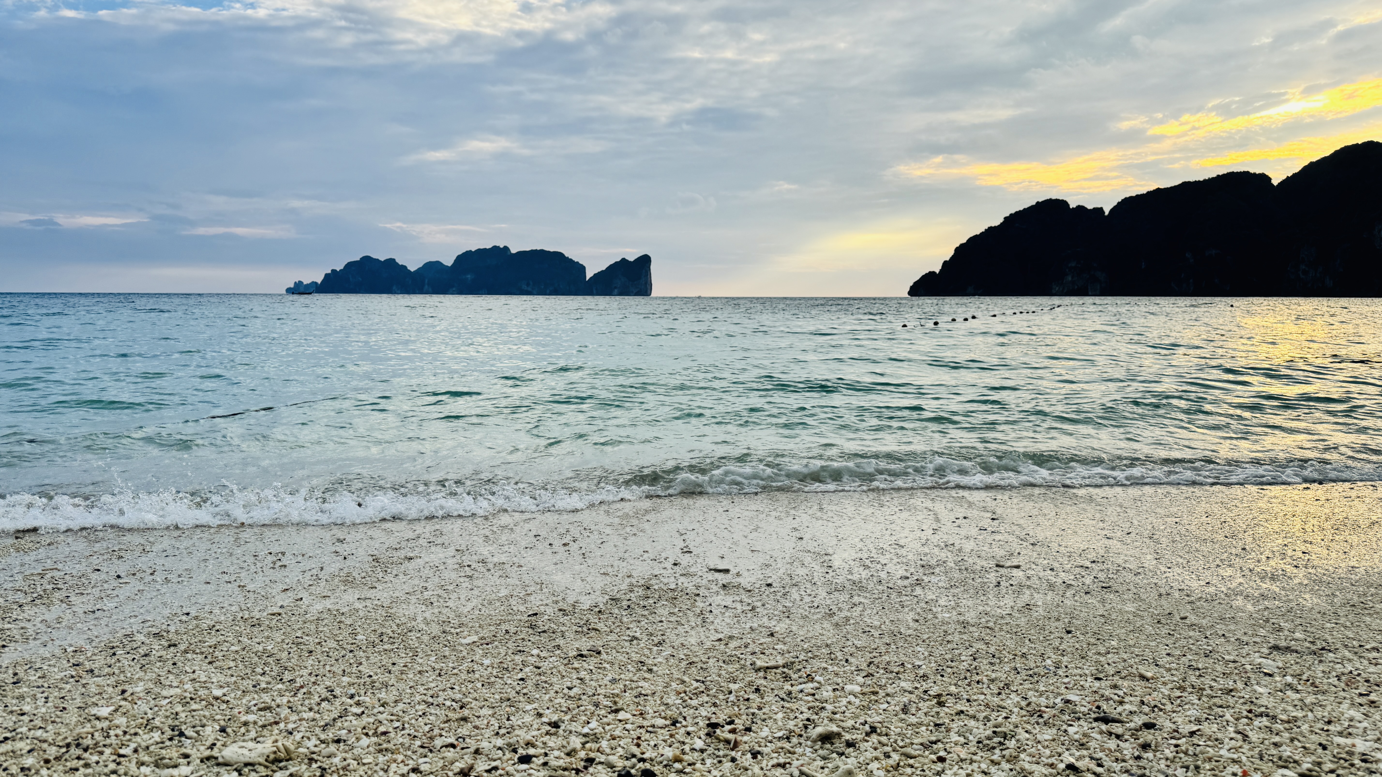 A wide view of a tropical beach at sunset, with pale sand, calm waves, distant rocky islands, and a warm glow under a cloudy sky.