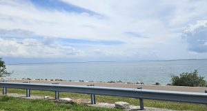 A serene view of the sea beneath a cloud-filled sky, with a distant bridge stretching across the horizon and a tiny city barely visible far beyond
