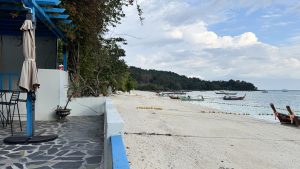 A view from a stone patio shows a long white sand beach and the ocean. There is a blue wooden structure with orange flowers on the left. Yellow buoys float in the water to mark a swimming area.